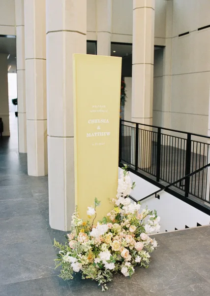 Wedding welcome sign, acrylic wedding welcome sign on a stand with white roses and greenery at a stone-columned indoor entryway staircase