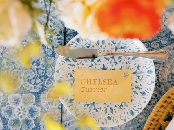 Wedding place card with calligraphy on a patterned blue and white plate, set on blue patterned linen with a wicker charger and butter knife