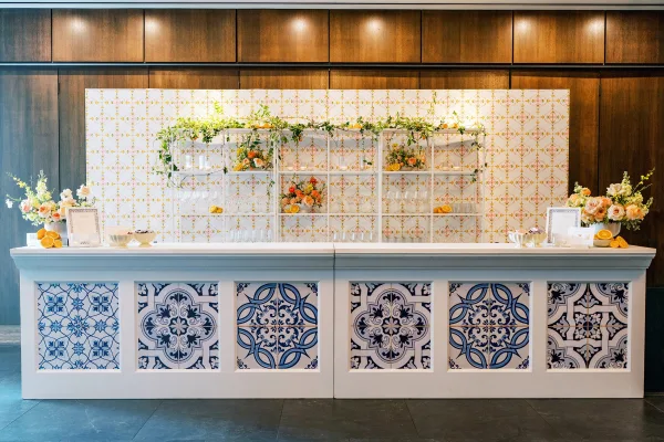 Wedding bar setup with a wedding cocktail bar counter, blue-and-white tile panels, glassware, florals, and citrus on shelves against wood walls