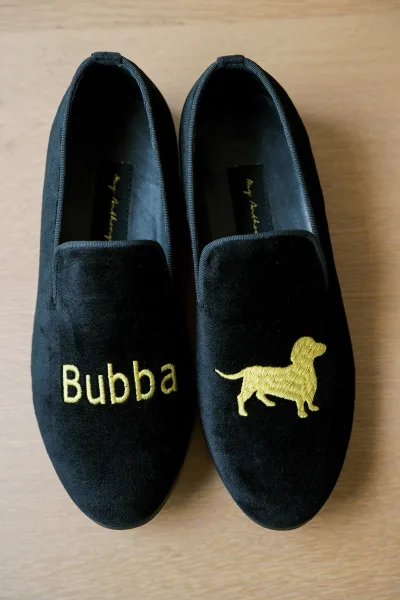 Groom wedding shoes in black velvet loafers with gold monogram and dog embroidery, displayed on a wood surface for getting-ready details