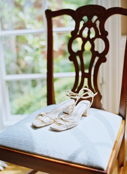Bridal shoes, white strappy heeled sandals with ankle straps on a carved wooden chair cushion by a window with garden greenery