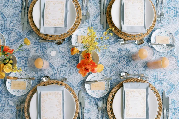 Reception tablescape with a blue and white tablescape on a blue patterned tablecloth, rattan chargers, orange blooms, lemons, and votives from above