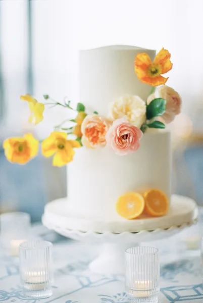 Wedding cake with white fondant, sugar flowers and orange slices on a cake stand, framed by votive candles on a white tablecloth