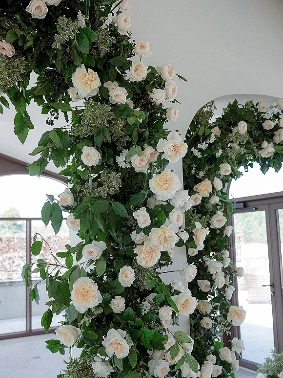 Wedding floral arch with white rose accents and cascading greenery garland framing arched windows and glass doors indoors