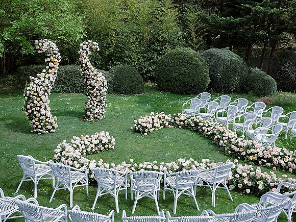 Ceremony setup for an outdoor wedding ceremony with white wicker chairs in a circle and a rose floral arch on a garden lawn
