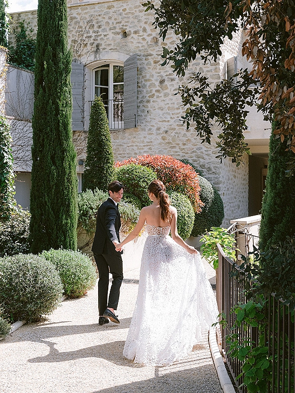 Couple portrait of bride and groom holding hands, bride in a strapless gown, walking by a sunlit stone villa and cypress trees.