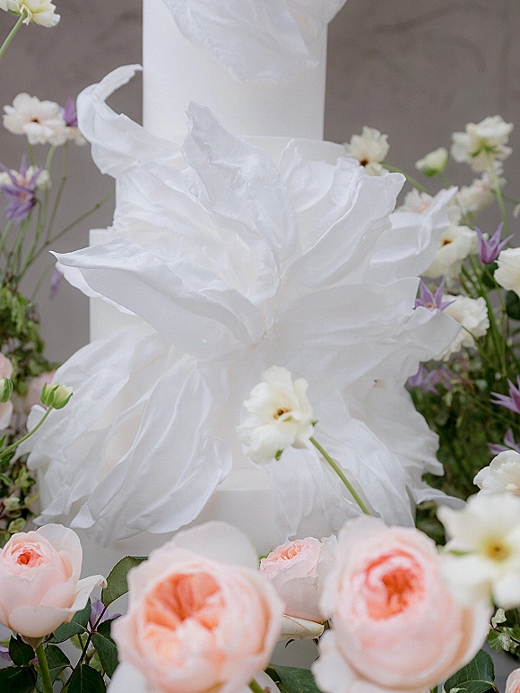 Wedding cake with white ruffle texture, adorned with fresh roses, white and purple flowers, and greenery against a neutral backdrop