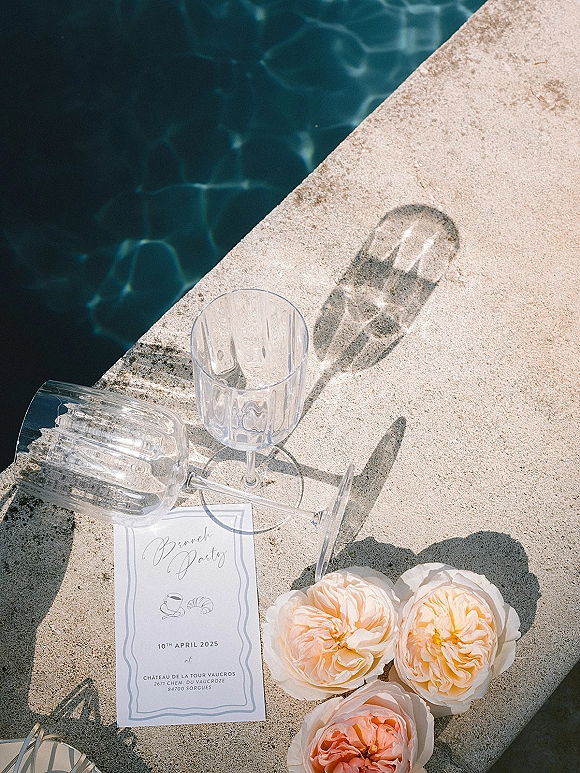 Wedding invitation with wedding stationery flat lay styling beside clear wine glasses and peach roses on a sunlit stone pool deck by blue water