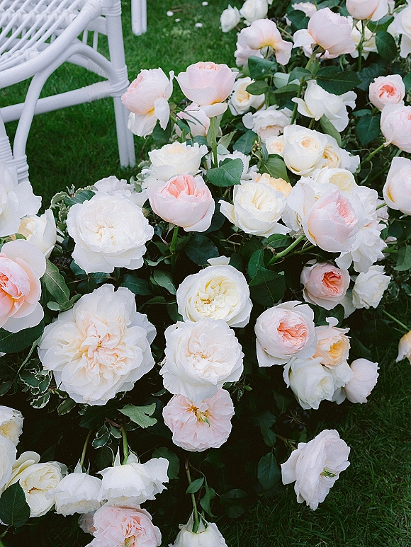 Wedding ceremony flowers with white and blush roses and greenery arranged on grass beside a white ceremony chair along the aisle