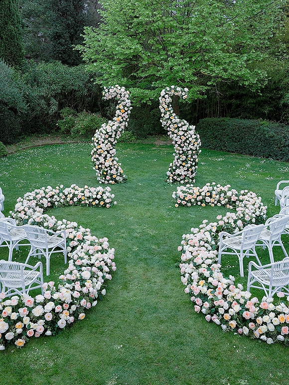 Ceremony aisle decor with outdoor ceremony aisle flowers lining a garden lawn, leading to floral arches framed by white chairs and trees