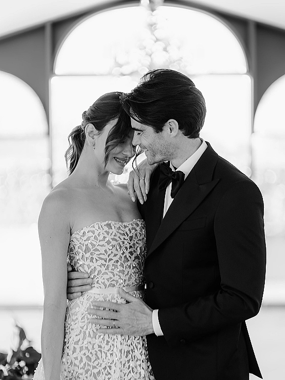 Couple portrait of bride and groom embrace with a forehead touch, bride in strapless lace dress by an arched window indoors