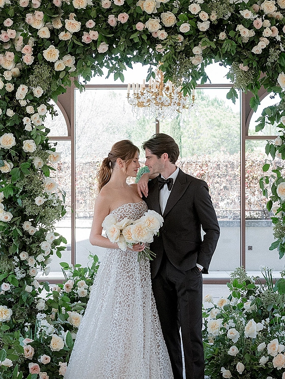 Couple portrait of bride in strapless lace wedding dress with white rose bouquet and groom in tuxedo beneath floral arch by arched windows