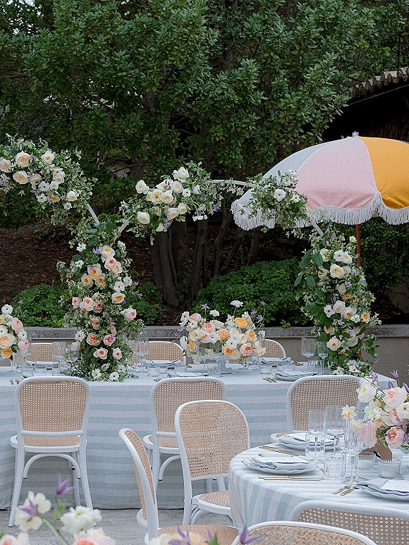 Reception tablescape at an outdoor wedding reception with striped linens, pastel centerpieces, greenery garlands, candles and a floral arch on a patio