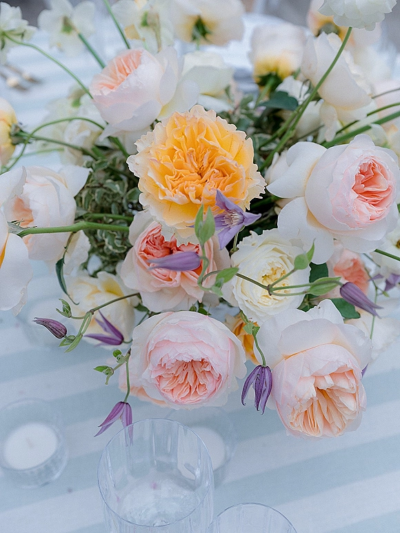 Wedding centerpiece flowers with peach and white garden roses, purple clematis, and greenery on white linen table with water glasses