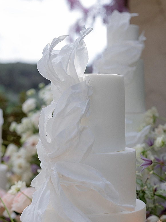 Wedding cake with white tiered wedding cake styling, sugar ruffle details and pastel flowers on a table outdoors with greenery and hills