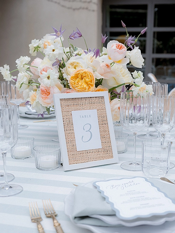 Reception tablescape with wedding table centerpiece of pastel roses and ranunculus, rattan-framed number, candles, striped linens by patio windows