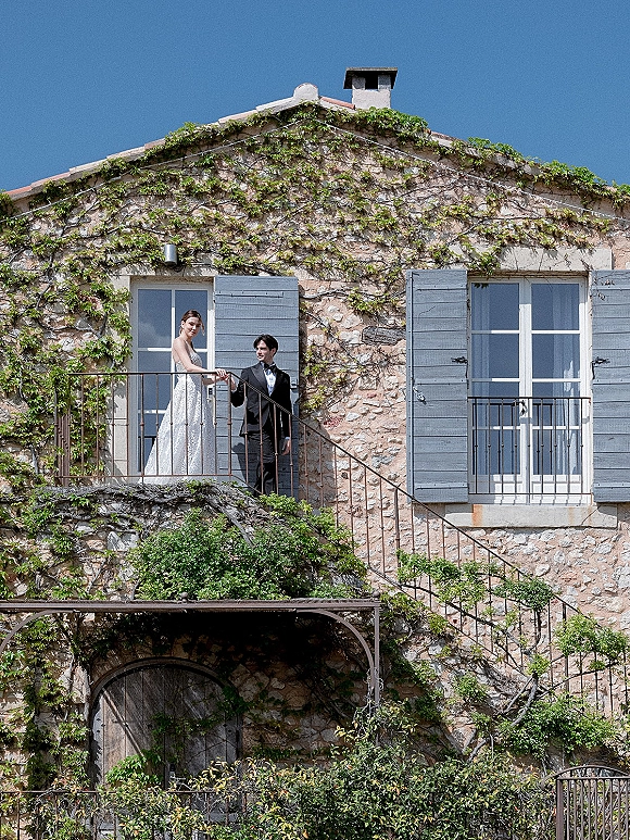 Couple portrait of bride and groom on balcony, holding hands by railing, framed by ivy on stone house with blue shutters and doors