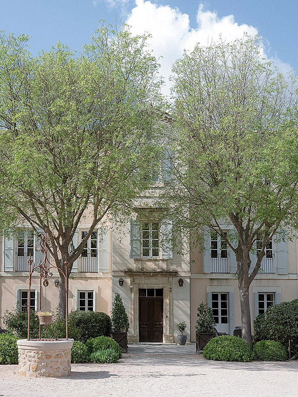 Wedding venue exterior with cream stucco facade, white shutters, double doors, and stone well in a gravel courtyard under blue sky