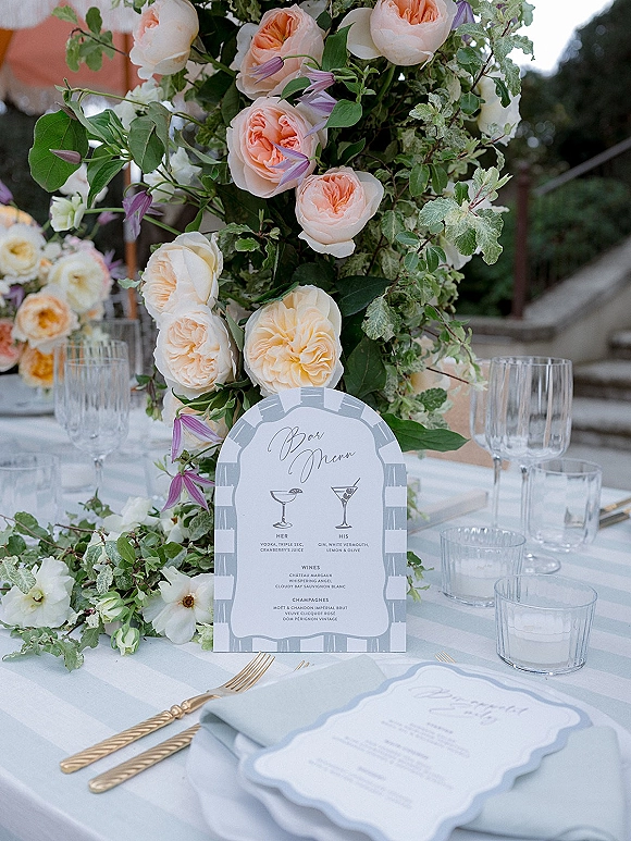 Wedding tablescape with reception table centerpiece of garden roses and greenery, gold flatware and menu cards on a striped cloth on an outdoor terrace