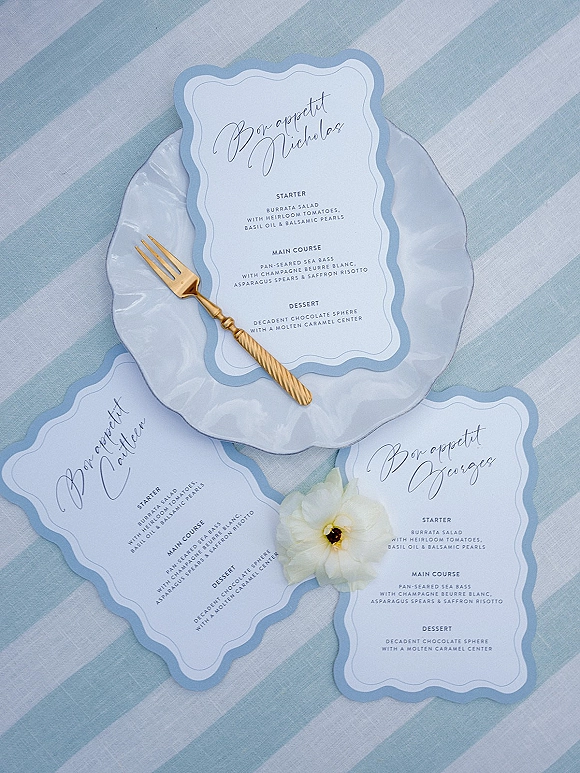 Wedding menu cards with calligraphy and scalloped edges rest on white plates with gold forks and a white flower on a blue striped tablecloth