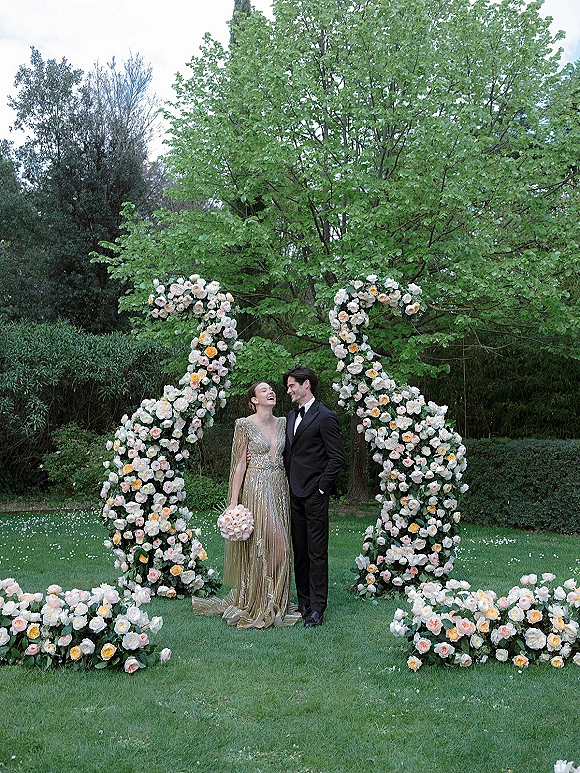 Couple portrait of bride in a beaded gown and groom in a tuxedo laughing under a rose floral arch on a garden lawn