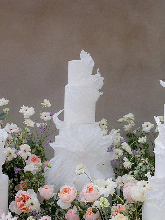 Wedding cake with white tiered wedding cake design, ruffled wafer paper and fresh roses, wildflowers, and greenery against a neutral wall backdrop