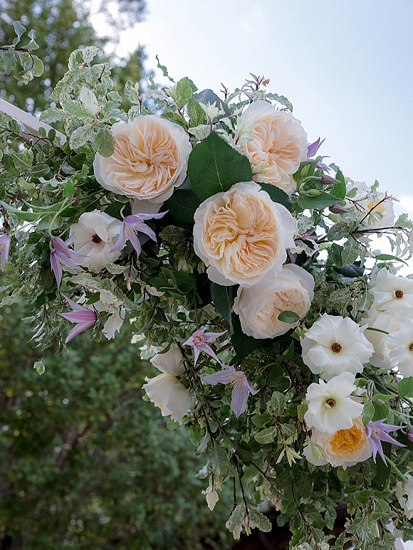 Floral arrangement on a ceremony arch with garden roses, white blooms, and lavender clematis accents against trees and sky