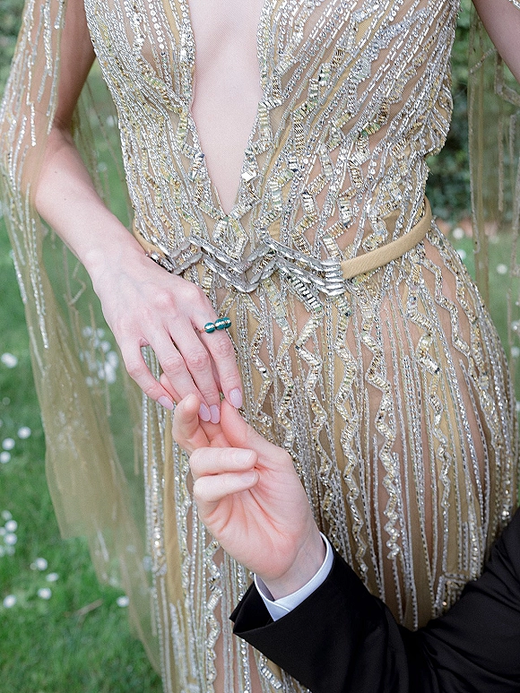 Engagement ring moment as groom holding bride hand, showcasing a turquoise ring against her beaded gown with garden foliage backdrop