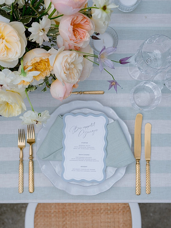 Reception tablescape with wedding place setting featuring peach rose centerpiece, gold flatware, scalloped menu card, and striped tablecloth background