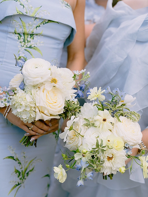 Bridesmaid bouquets of white and blue bridal bouquet blooms with greenery, held by manicured hands in pale blue dresses, softly blurred behind