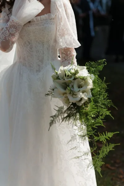 Bridal bouquet of white calla lilies with cascading greenery and fern fronds, held against a lace-sleeved dress with blurred guests outdoors