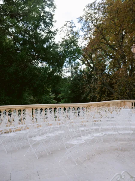 Ceremony seating with outdoor ceremony chairs in white iron, arranged on a stone terrace by a balustrade, framed by garden trees and sky.