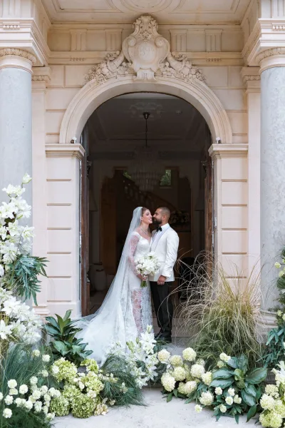 Couple portrait with the bride kissing groom, her veil drifting as they stand by stone columns and an arched doorway beneath a chandelier.