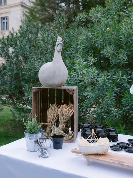 Appetizer station for a wedding, styled with a ceramic chicken statue, set beside garden shrubs and a stone building facade.