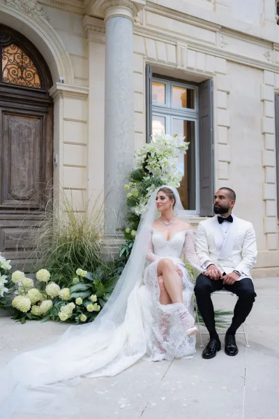 Couple portrait of bride and groom seated by a stone column and wooden door, highlighted by white florals as they share a quiet pose.