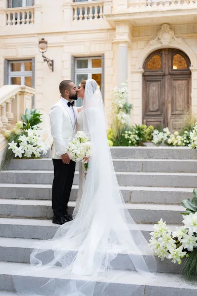 Wedding kiss portrait of a bride and groom on stone steps outside a historic building, her long veil trailing as they embrace before wooden doors.