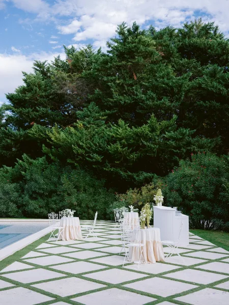 Cocktail hour setup features outdoor cocktail seating with white bistro chairs on a checkerboard patio beside a serene swimming pool.