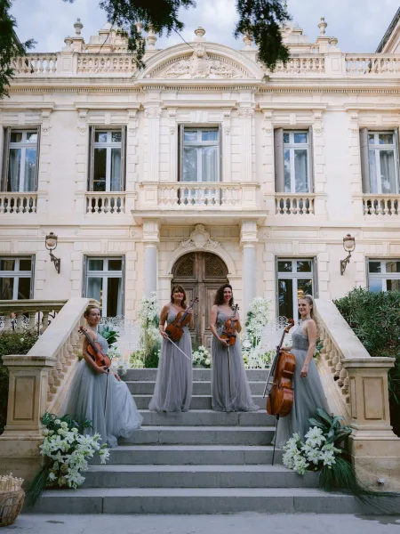 Wedding string quartet serving as ceremony musicians stands on a stone staircase at a historic mansion, wearing elegant gray gowns.