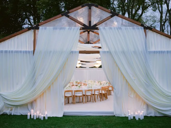 Tented wedding reception inside a clear tent reception, chandeliers glowing as dusk settles over a lawn with trees and a soft evening sky.