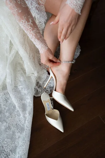 Bridal shoes—ivory wedding heels with a crystal ankle strap, resting on a dark wood floor, their subtle sparkle contrasting the rich grain.