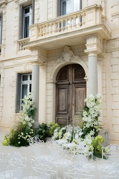 Outdoor ceremony setup with wrought iron wedding chairs facing arched wooden doors on a stone facade, framed by columns and a balcony