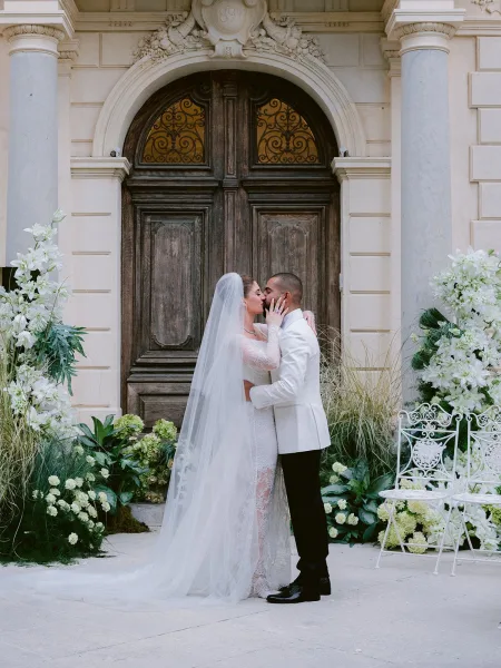 Wedding kiss portrait as the bride and groom embrace beneath ornate wooden doors and a stone archway, her lace wedding dress softly flowing.