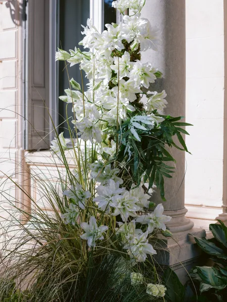 Floral installation creates a lush floral column wrap with white lilies climbing a stone column against a building facade and garden foliage.