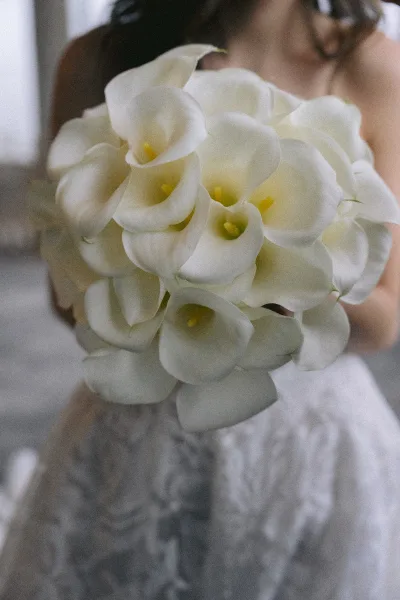 Bridal bouquet of white calla lilies held close, partially hiding the bride’s face, with lace gown details against a softly blurred interior backdrop