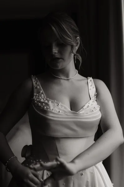 Bridal portrait of a bride by window in black and white, wearing a pearl-embellished dress and pearl necklace beside soft curtains indoors