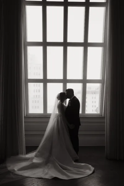 Wedding kiss portrait of a bride and groom silhouette by a large window, her long veil and train flowing as city buildings glow outside
