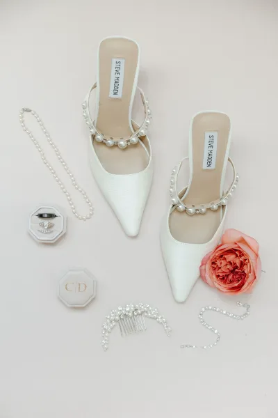 Bridal shoes flatlay featuring white wedding heels with pearl straps, rings and jewelry beside a coral rose on a blush background surface