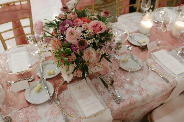 Reception tablescape with wedding table centerpiece of blush roses, ranunculus, and greenery, set with gold-rim chargers, menus, and votive candles in a reception room