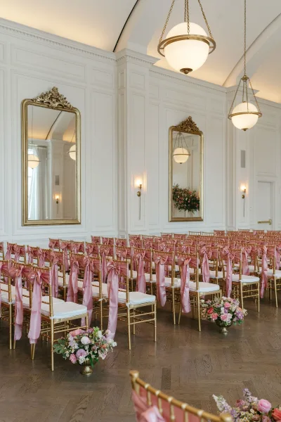 Ceremony seating with gold chiavari chairs lined in rows, pink chair bows, and floral aisle arrangements beneath globe pendant lights in a white paneled ballroom