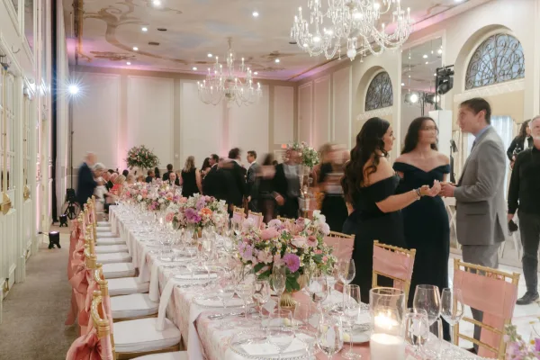 Reception tablescape with long banquet head table, blush floral centerpieces, taper and hurricane candles, gold Chiavari chairs under chandeliers in a ballroom
